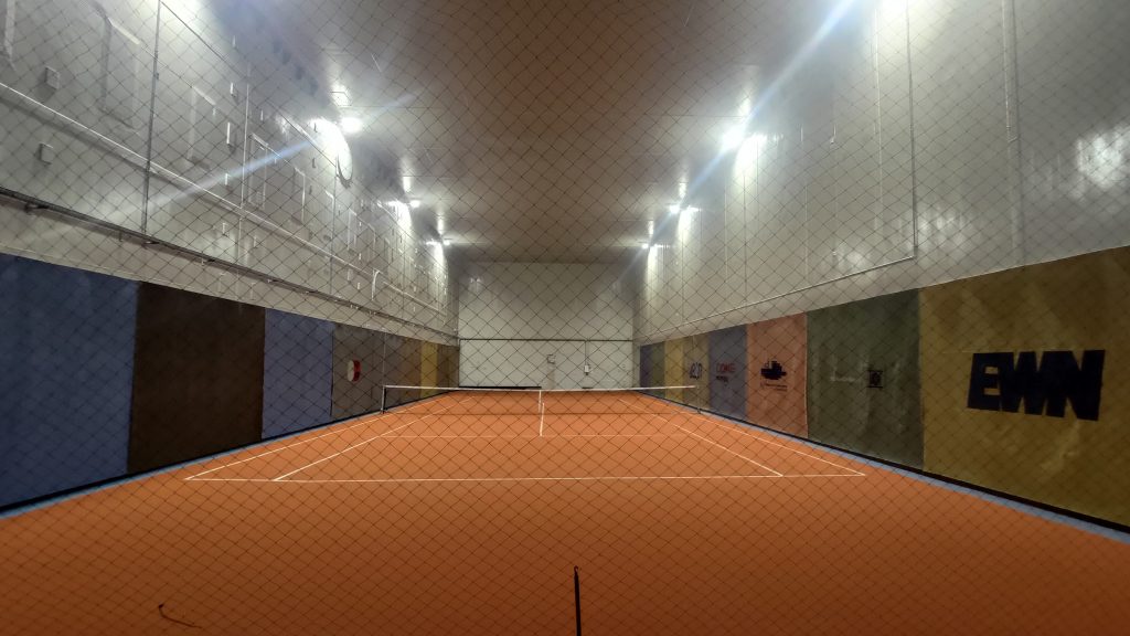 Ein Tennisplatz in einem hohen, mit Stahlplatten beschlagenen Raum, fotografiert durch ein Netz.