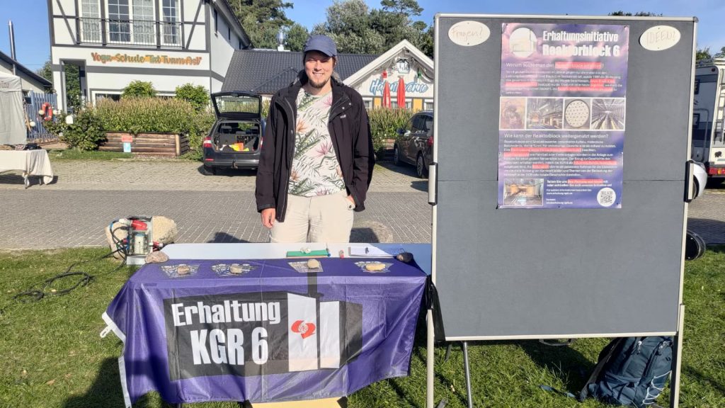 Ein Mann steht hinter einem Tisch auf dem Flyer und eine Flagge mit dem Logo der Erhaltugnsinitiative liegen. Neben dem Tisch steht eine Pinnwand mit einem Plakat der Initiative.