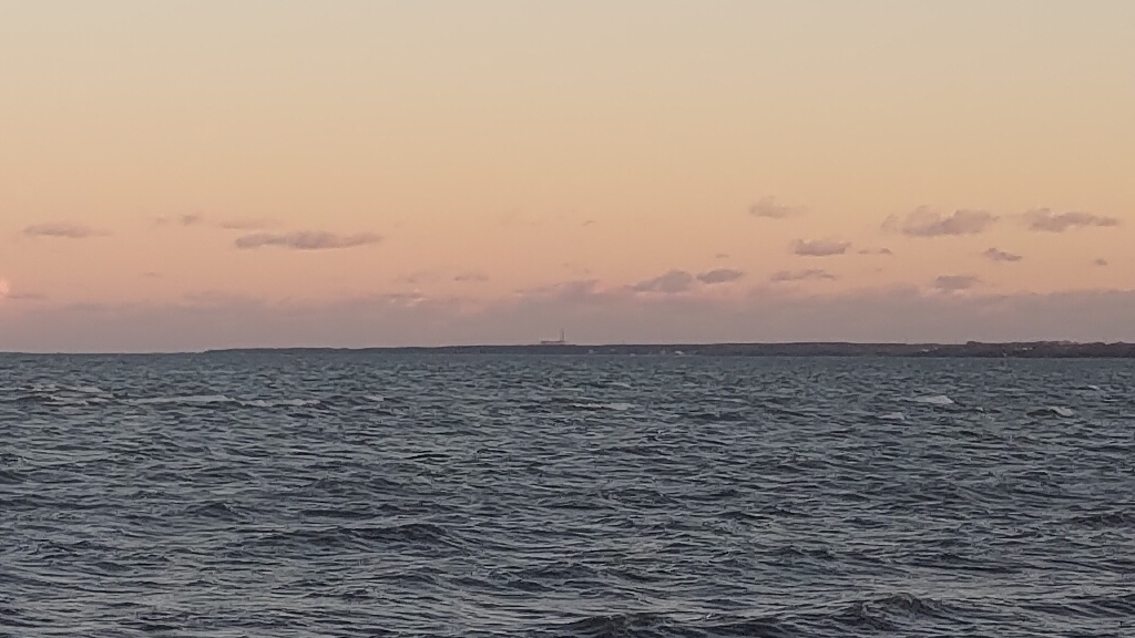 Blick über leicht gewellten Bodden auf die Silhouette des KKW Lubmin vor einem leicht orangen Abendhimmel.